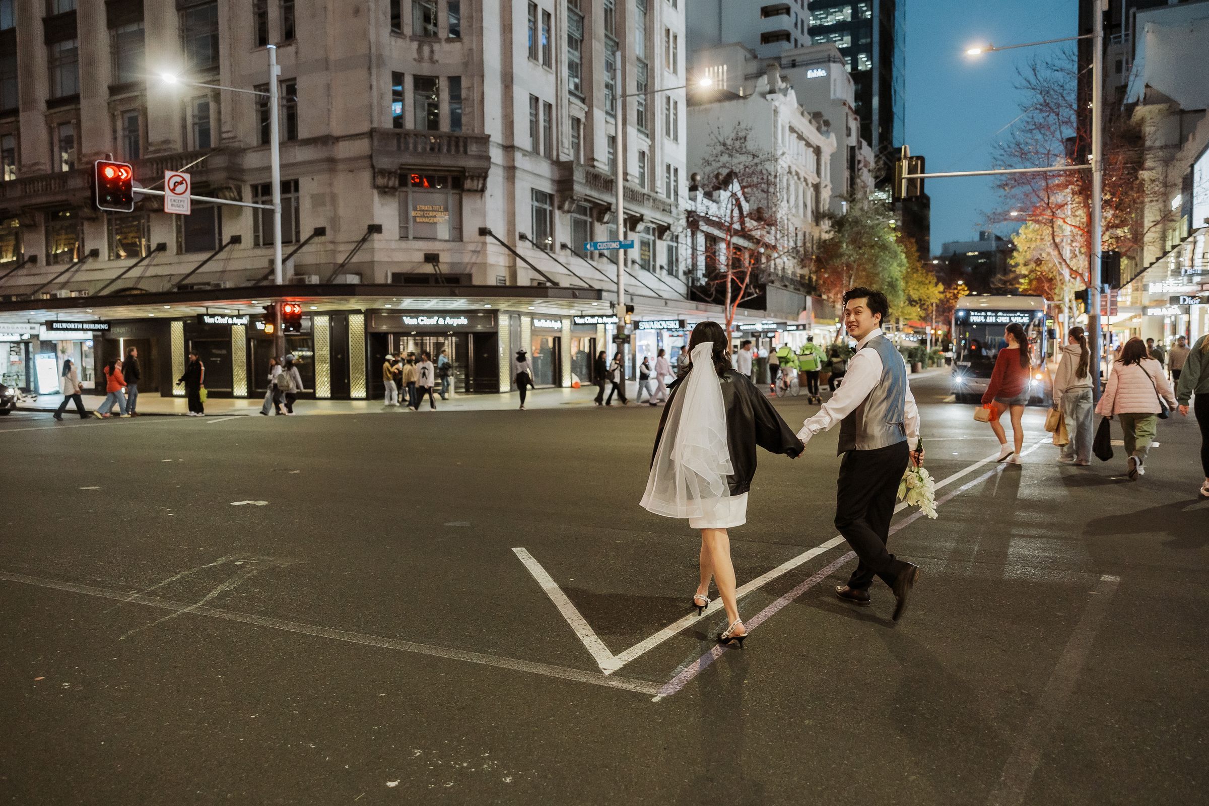 Bride and groom crossing Queen Street in Auckland at night with city lights behind